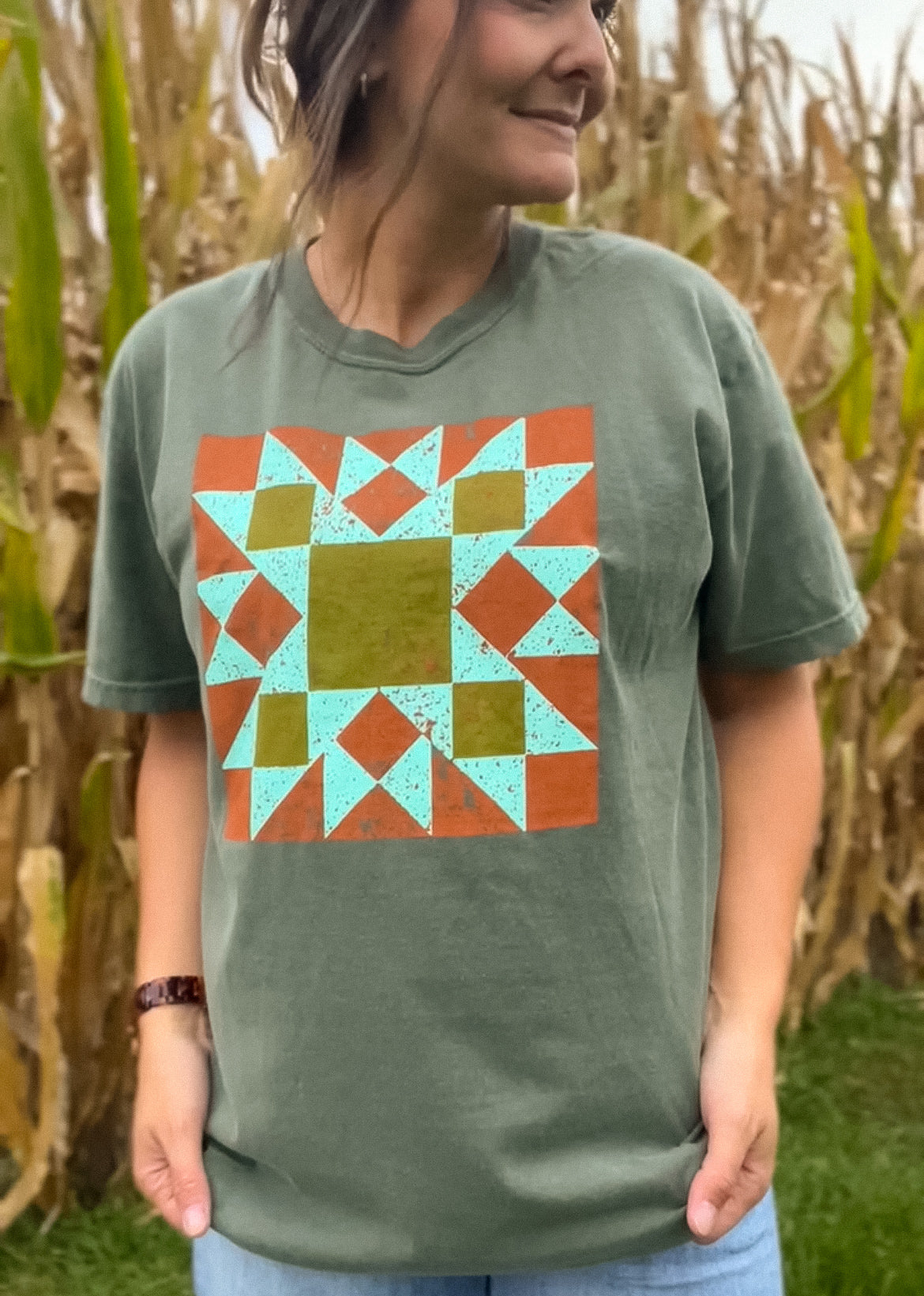 A woman wearing an olive green t-shirt with a colorful geometric barn quilt pattern graphic on the front, standing in a cornfield.