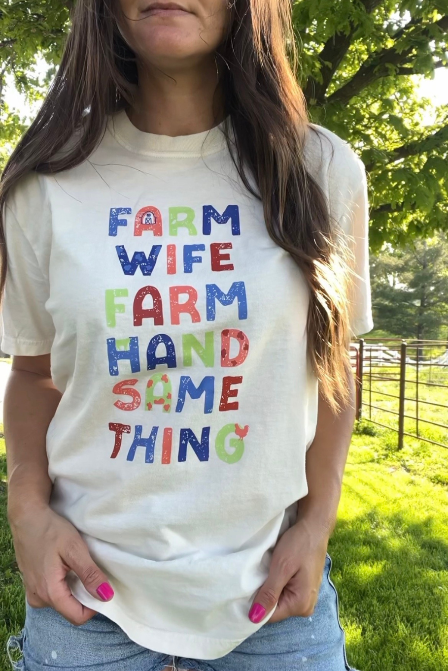 Woman wearing a white tee with a multicolored graphic saying "farm wife, farm hand, same thing" on the front of the shirt.