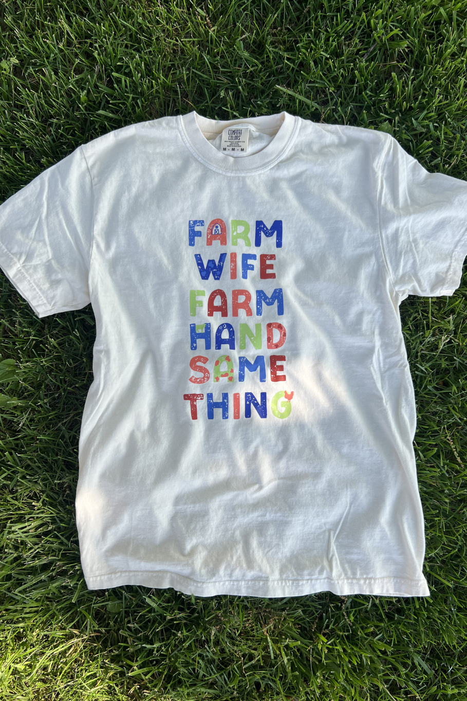 White tee with multicolored "farm wife, farm hand, same thing" graphic on front of shirt.
