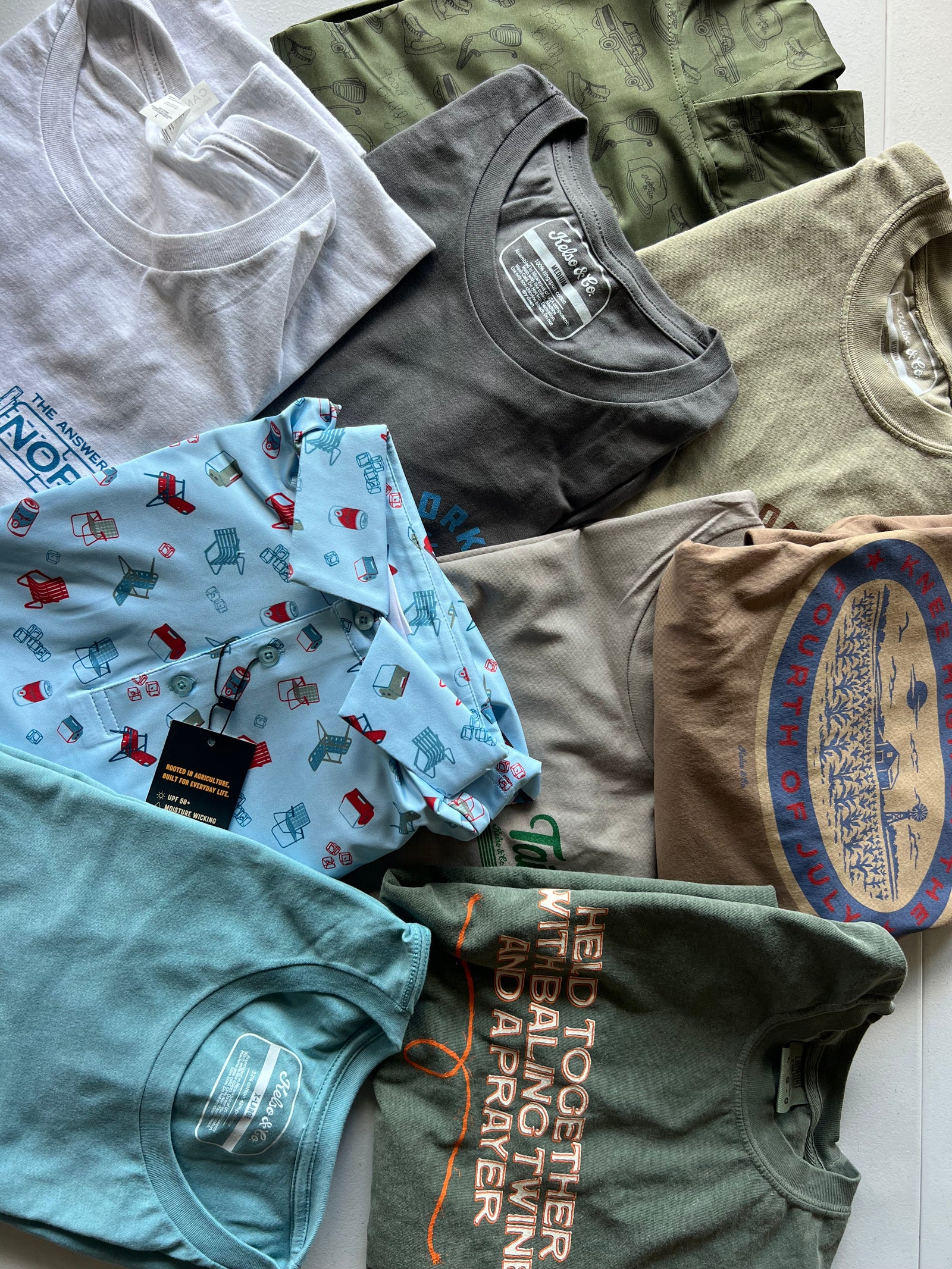 A flat lay of multiple folded graphic t-shirts in various colors and patterns, including light blue, gray, olive green, and beige, arranged on a white floor.