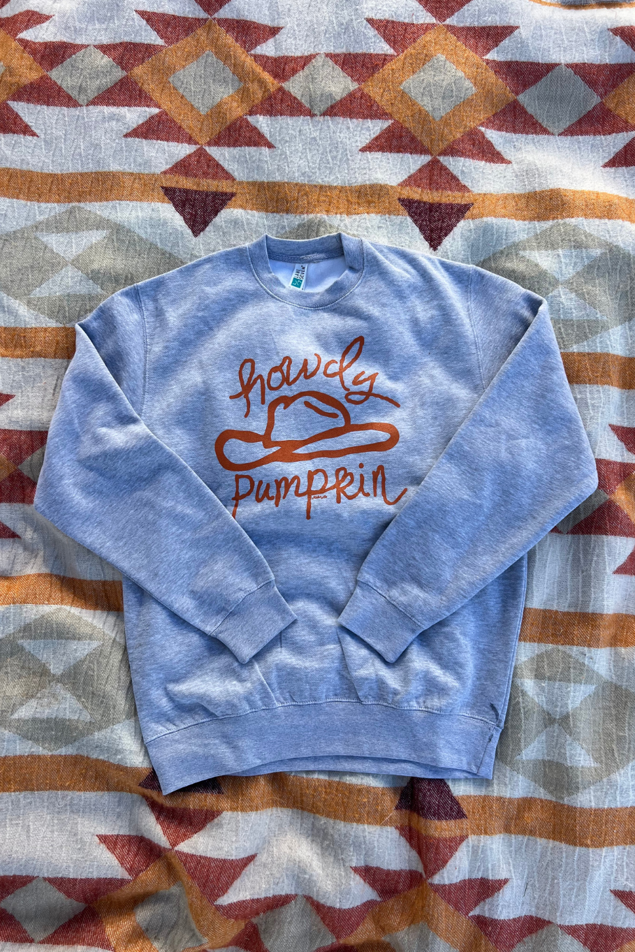 A light gray heather crewneck sweatshirt with a burnt orange graphic reading 'howdy pumpkin' and a cowboy hat outline, laid flat on a patterned western blanket.
