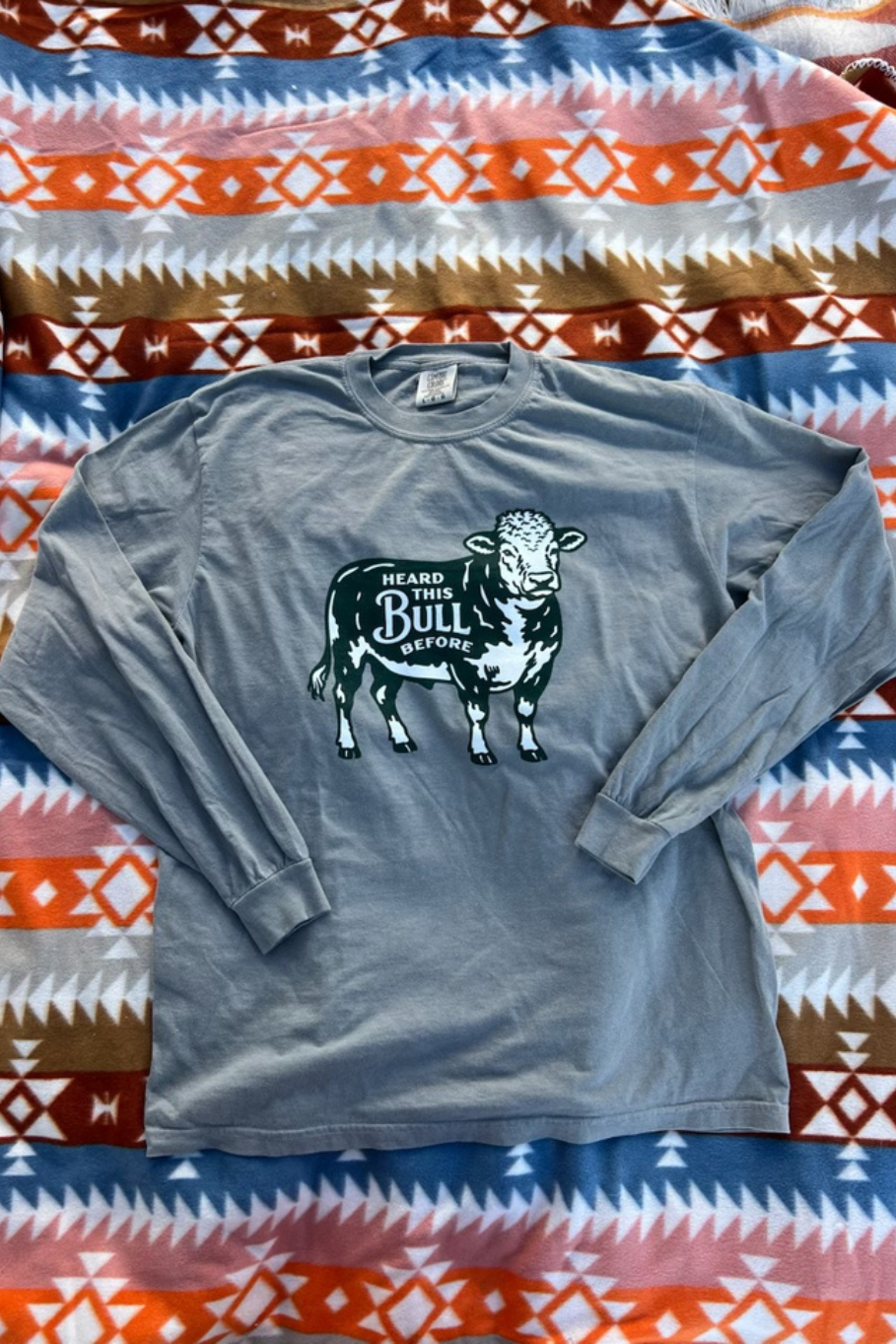 A gray long sleeve with a bull graphic on the front. It says "heard this bull before". Shirt is laying on a western printed quilt.