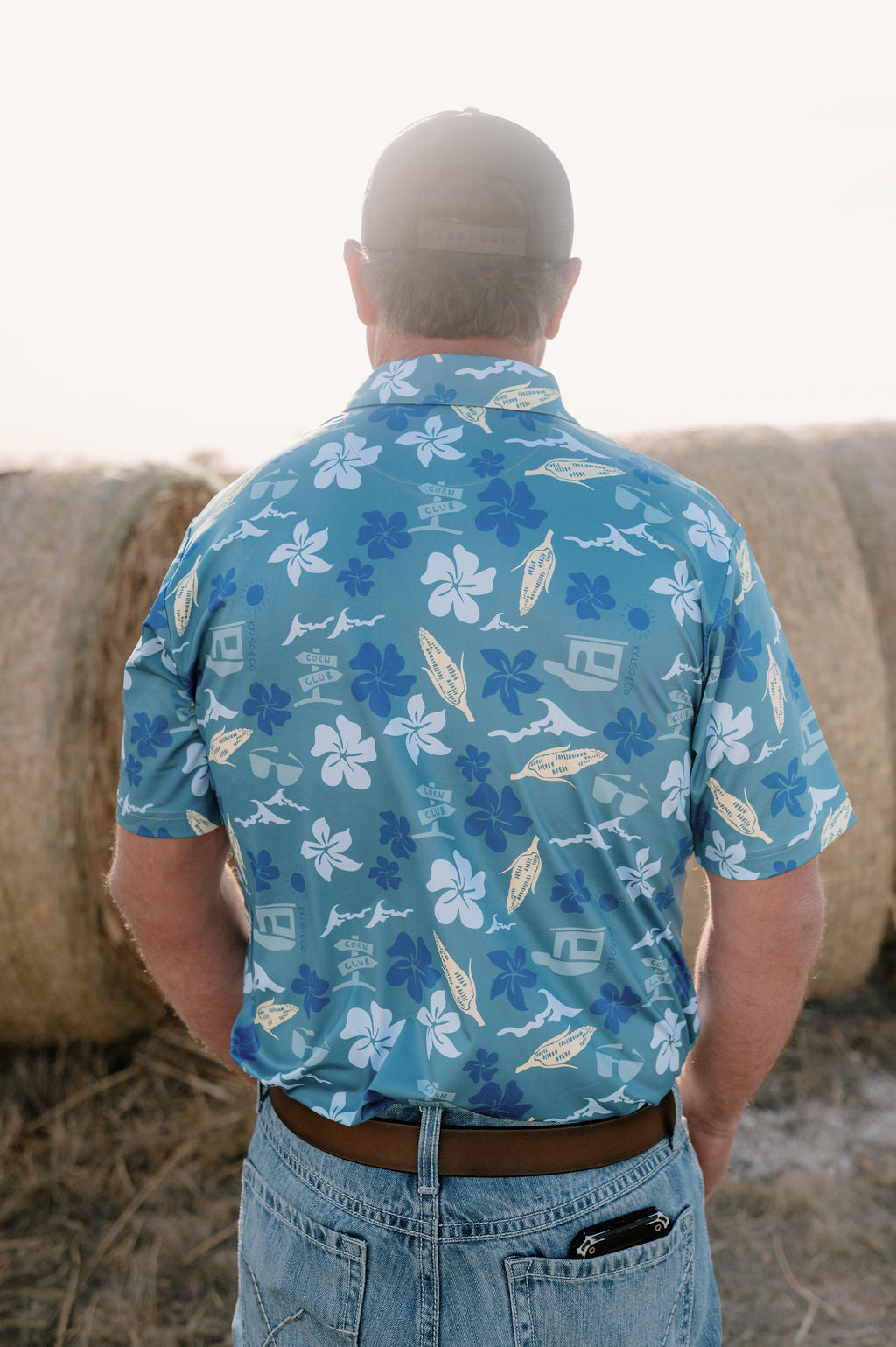 Picture of the back of a man standing in front of hay bales wearing a blue Hawaiian inspired polo.