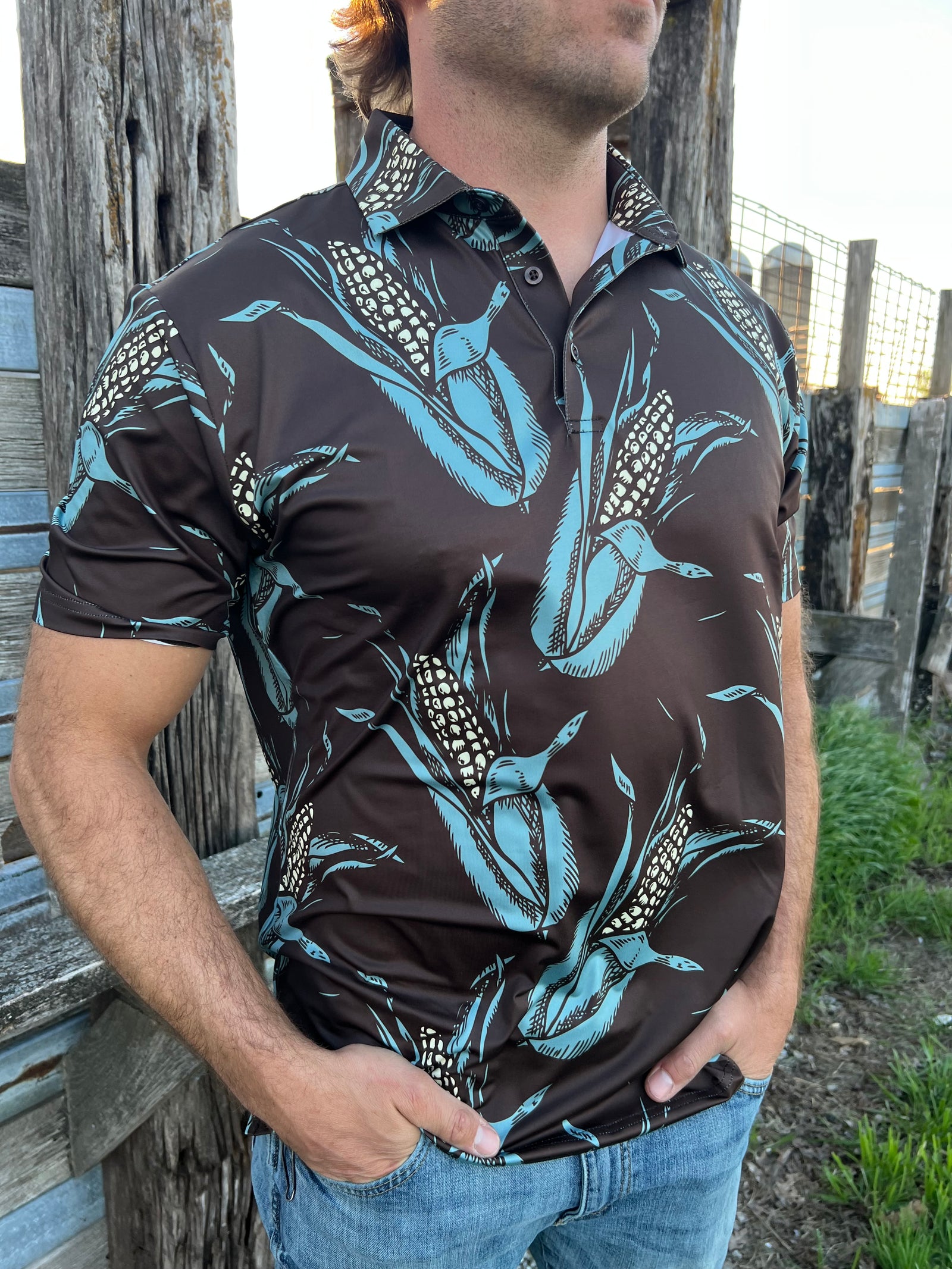 Man standing in front of a fence wearing a dark brown polo with ears of corn all over it.