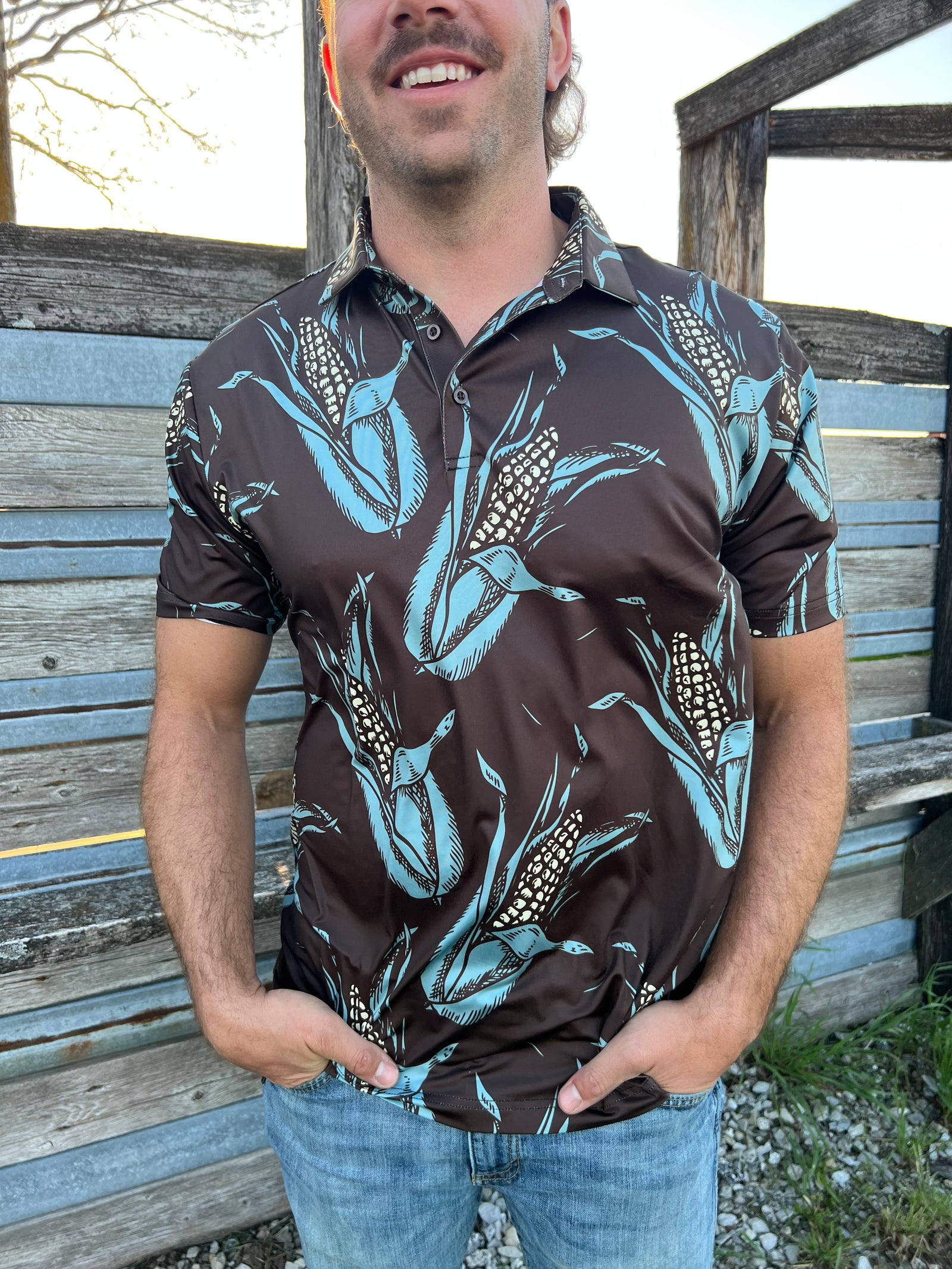 Man standing in front of a fence smiling, wearing a brown polo with ears of corn all over it.