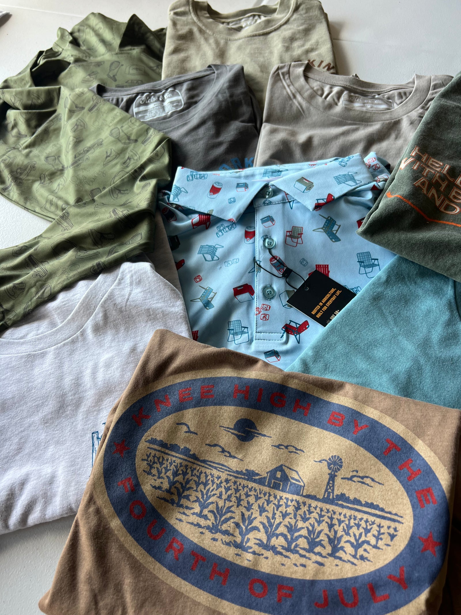 A flat-lay collection of folded graphic t-shirts and performance hoodies in various colors and patterns, including olive green, light blue, beige, white, and teal, with one prominent brown shirt featuring a blue and red 'Summer High by the Fourth of July' graphic.