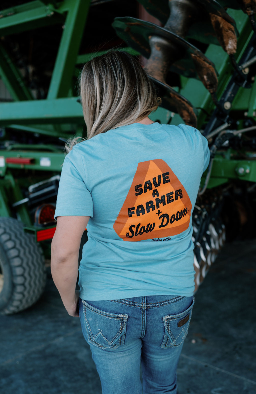 woman wearing a blue tshirt in front of a farm implement, the back has an orange slow moving vehicle sign and says "save a farmer + slow down"