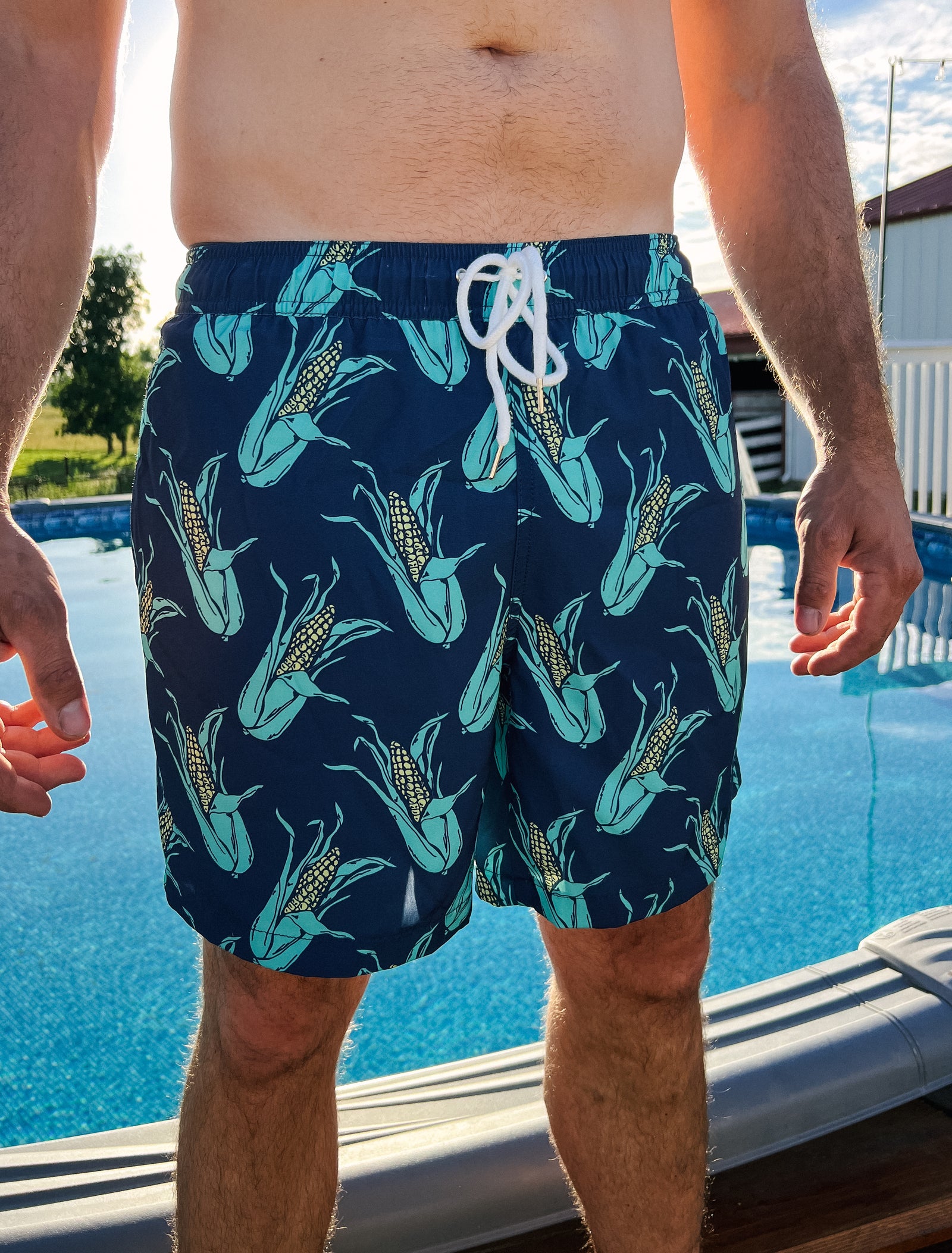 Man standing in front of pool wearing the shuck yeah swim trunks that are navy blue and have a corn print.
