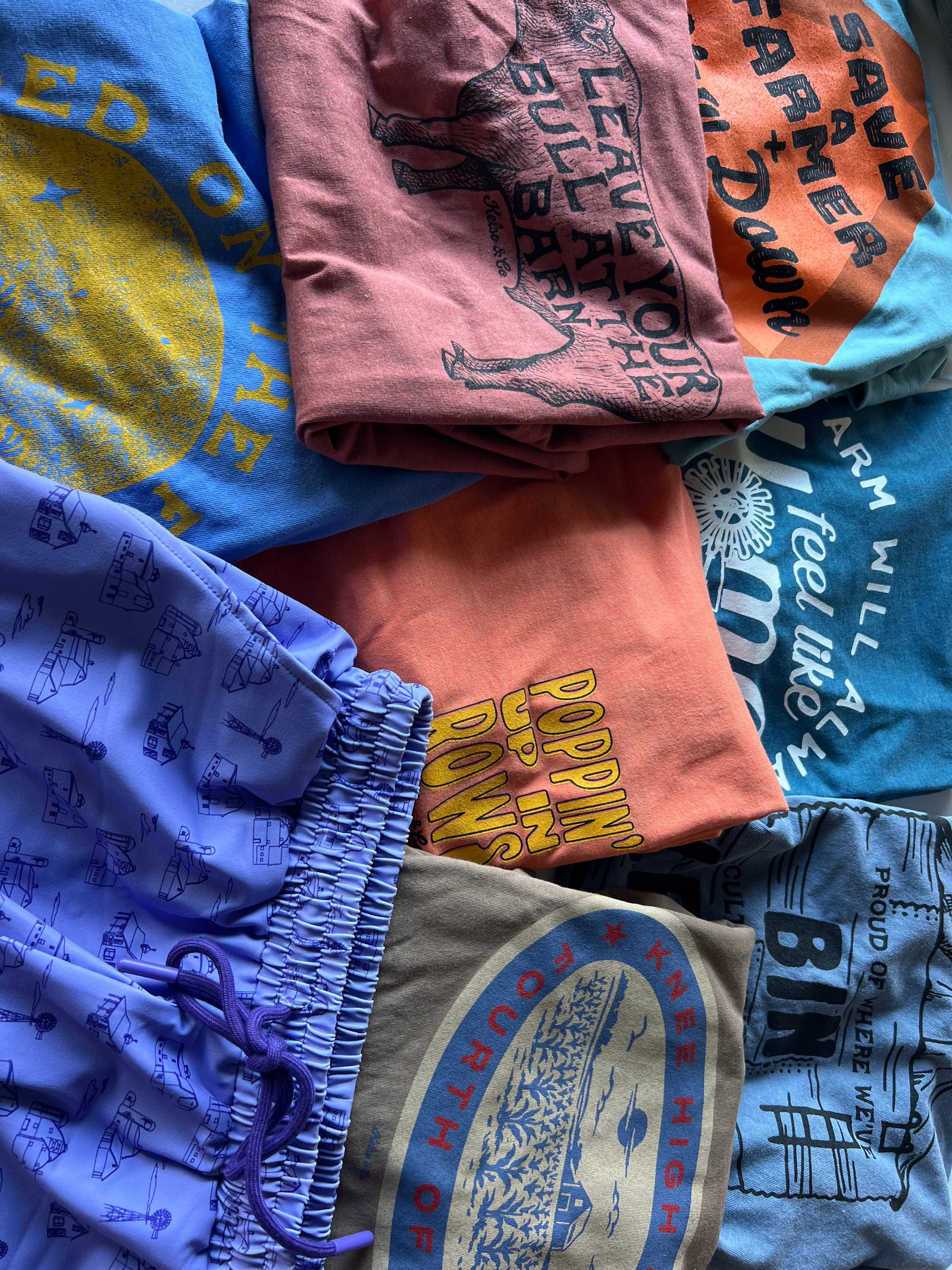 A pile of folded graphic t-shirts and shorts in assorted colors, including blue, pink, orange, purple, and gray, each featuring vintage-style printed graphics and text.