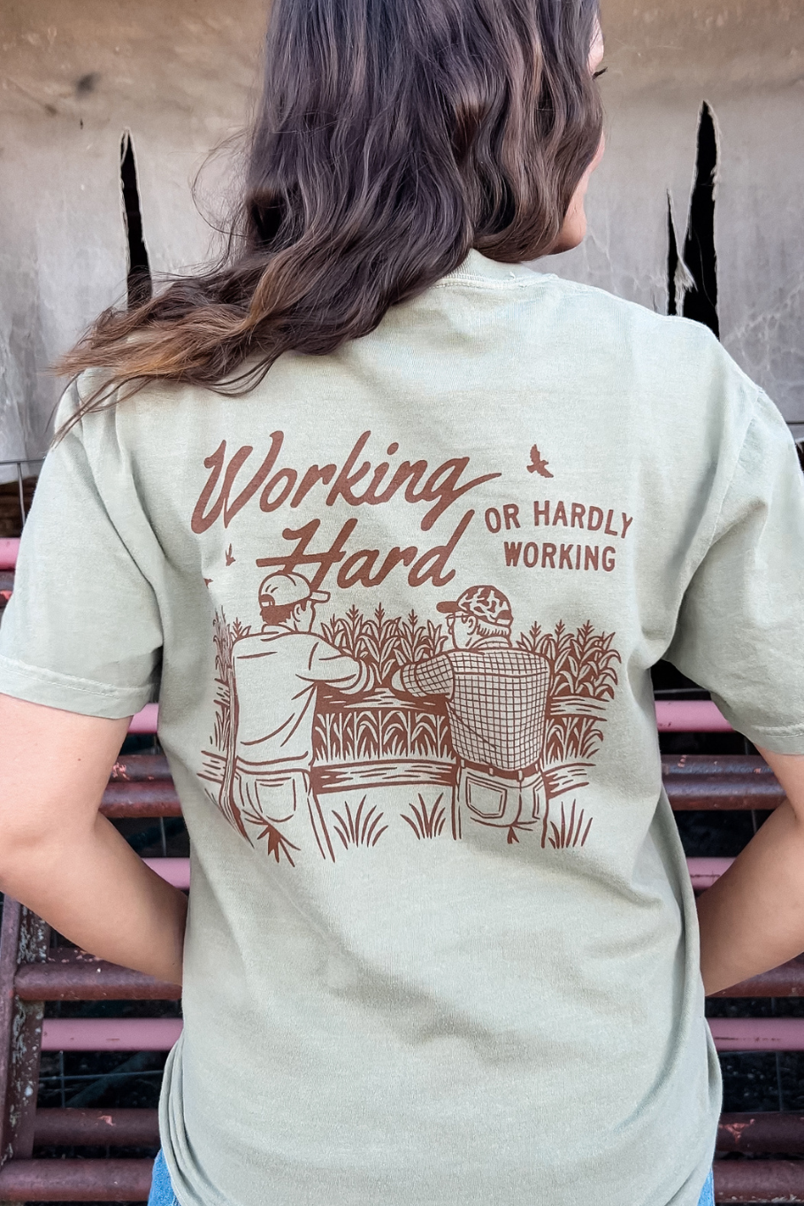back of woman wearing khaki shirt that has two farmers on it that says working hard or hardly working