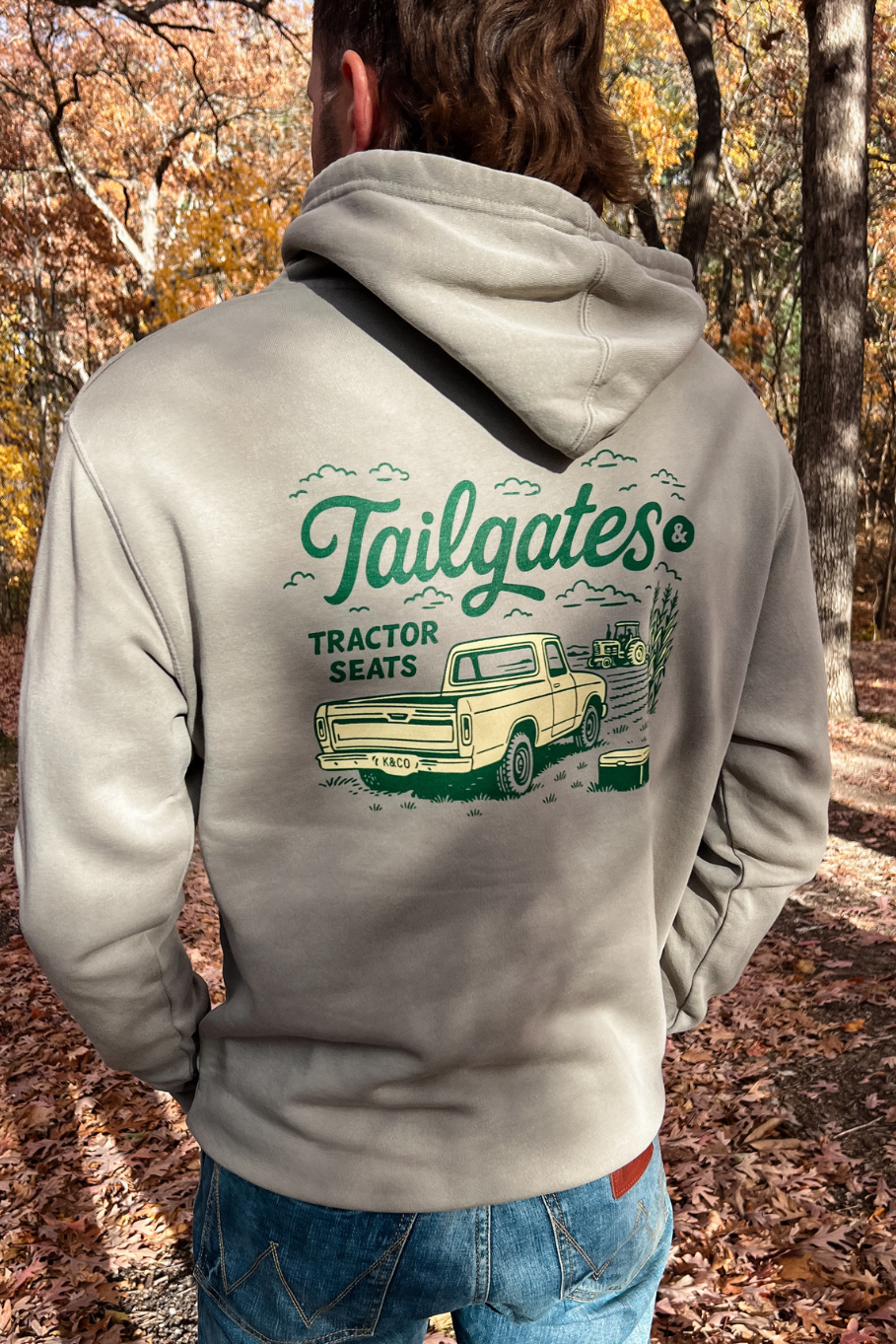 back of guy in a cement pigmented hoodie that says tailgates and tractor seats in green and gold with an old pick up truck and a tractor in the field