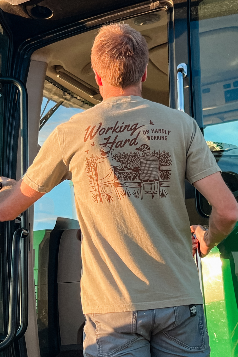 Guy climbing into a tractor with a khaki short sleeve shirt on that says working hard or hardly working