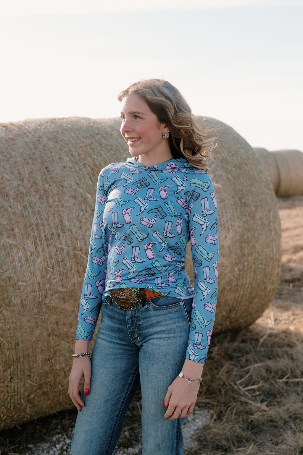 girl on a farm in front of hay bales with the boot scootin performance hoodie - a periwinkle base with multip colored muted cowgirl boots in greens, blues and purples