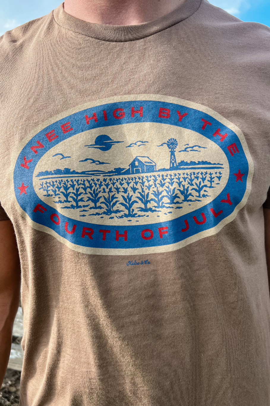 close up of a Guy with a light brown tshirt on and a graphic in blue, red and tan that says "knee high by the fourth of july." The graphic shows a corn field with a barn and windmill