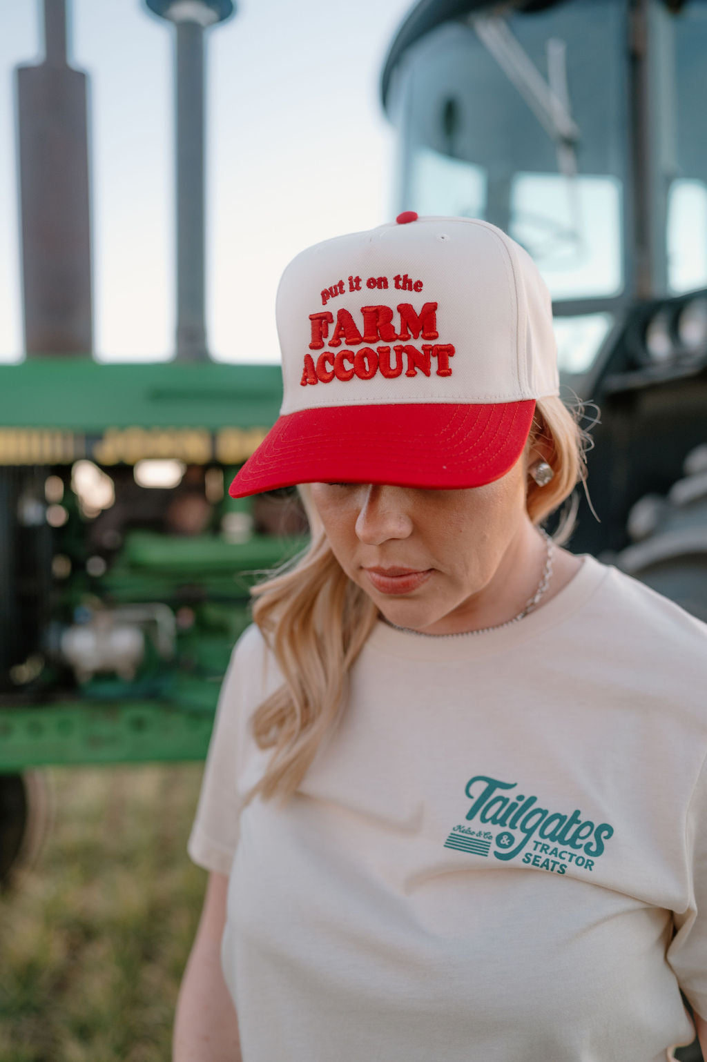 Red and natural trucker hat on girl standing in front of a tractor. Hat is embroidered with "Put it on the farm account"in red