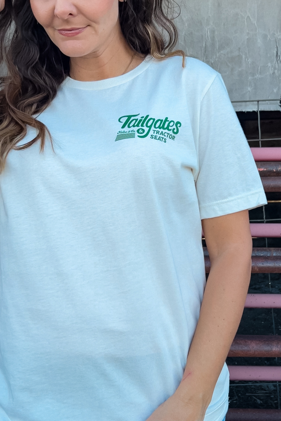girl standing in front of cattle panels showing the front of her t-shirt that says "tailgates & Tractor seats" on the left chest in green print