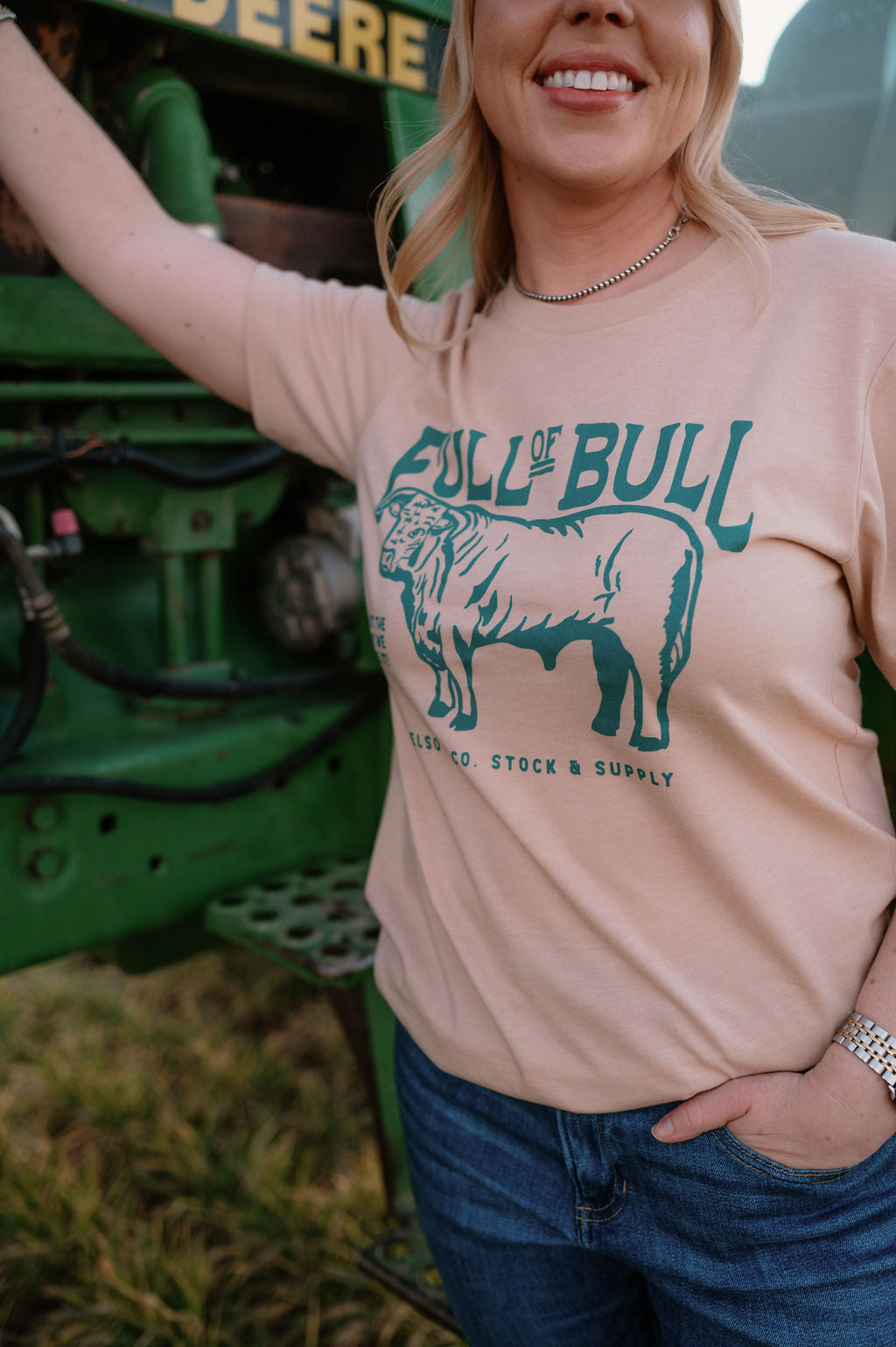 woman standing against a john deere tractor with a tshirt that says full of bull with a vintage illustration of a bull