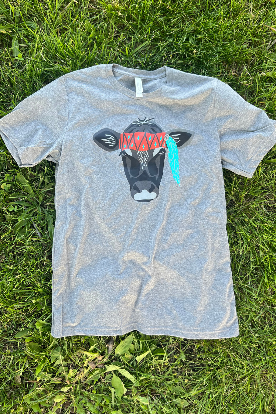 flatlay of a women's gray triblend graphic tee with a black cow face with a red band across her forehead and a feather hanging down from her left ear