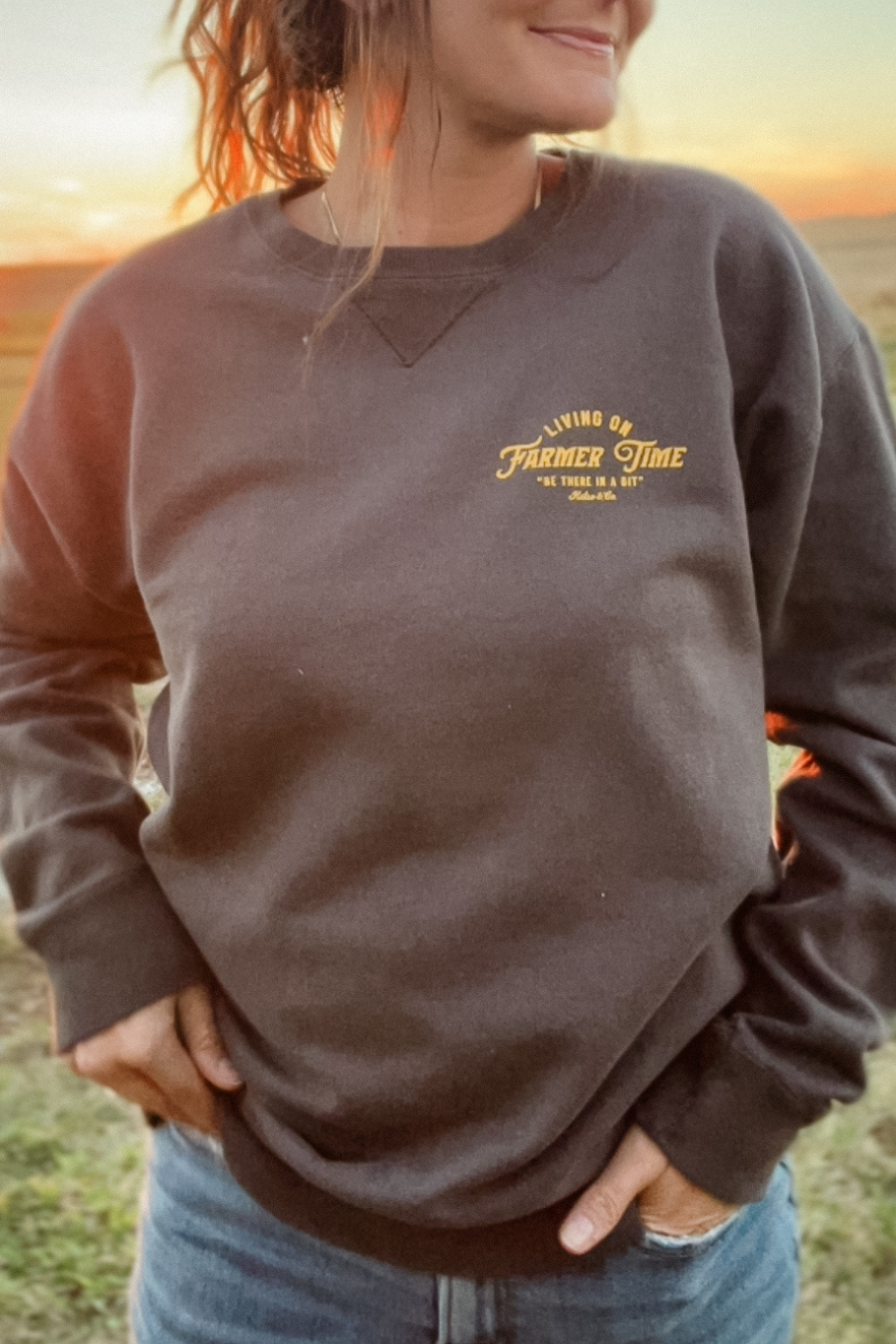 girl on a farm in front of a sunset wearing a crewneck that says Living on farmer time