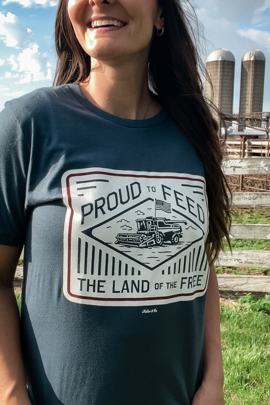 woman in front of a white fence with a blue graphic tshirt on that says "proud to feed the land of the free." The graphic has a combine with an american flag