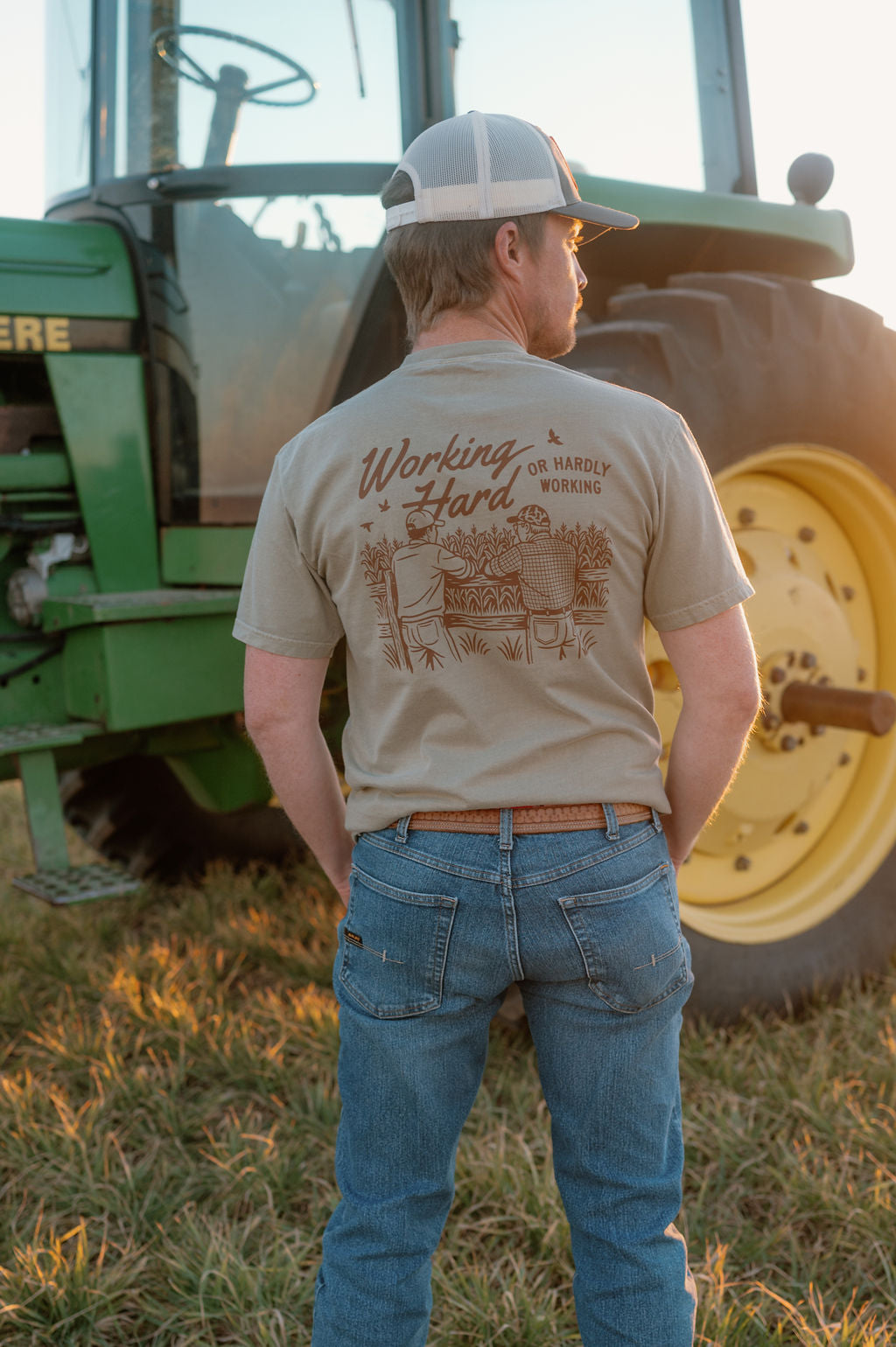 guy standing in front of a tractor wearing a graphic tshirt that says working hard or hardly working