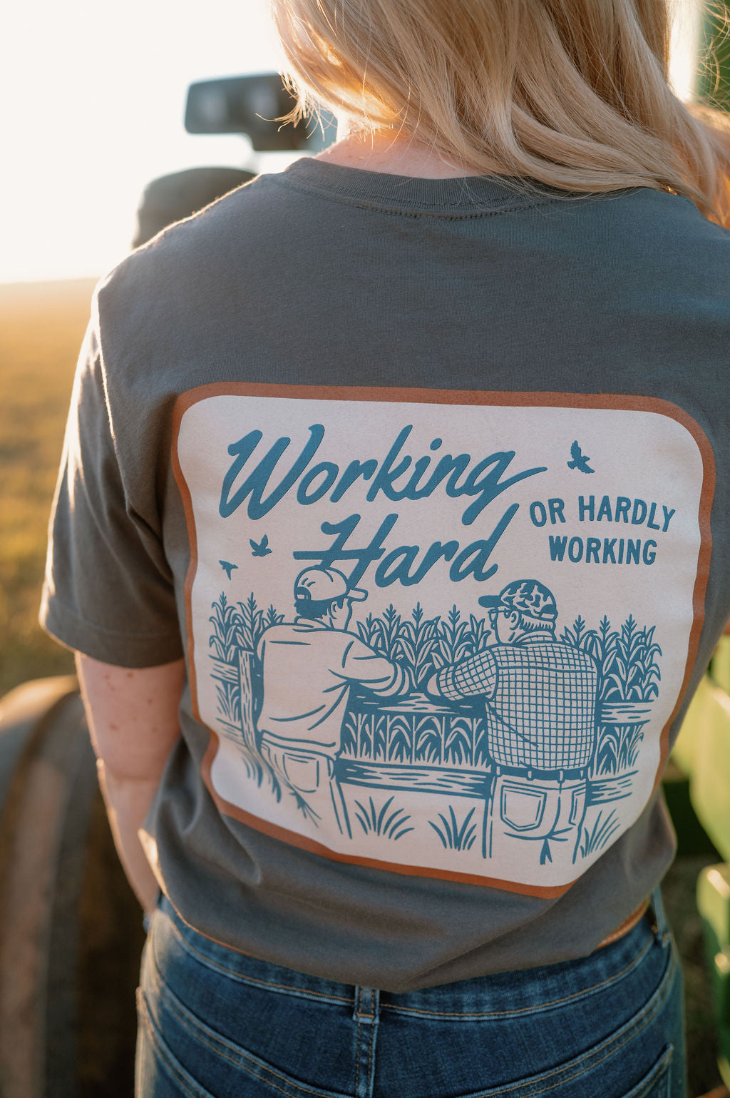 girl wearing a tshirt showing the back design of "working hard or hardly working" with two farmers leaning on a fence talking, looking out into a corn field