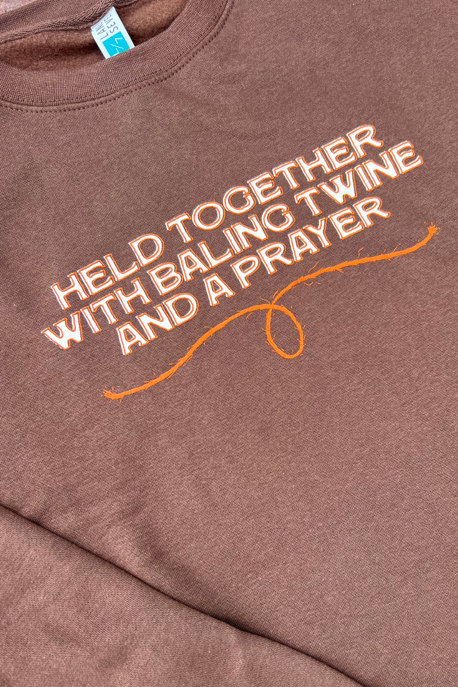 White and Orange "Held together with baling twine and a prayer" graphic on a brown crewneck, close up shot. 