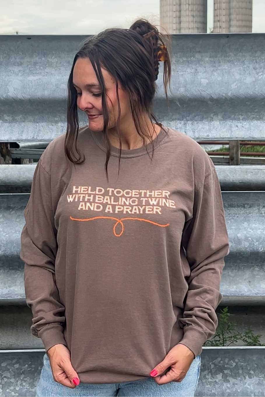 A woman wearing a brown long-sleeve graphic sweatshirt with the text 'HELD TOGETHER WITH BALING TWINE AND A PRAYER' in orange and cream, standing outdoors in front of corrugated metal steps.