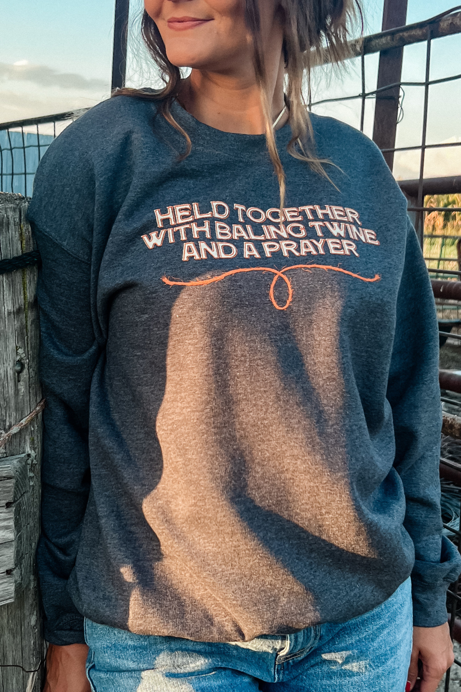Woman standing in front of a gate on a farm in the baling twine sweatshirt in dark gray