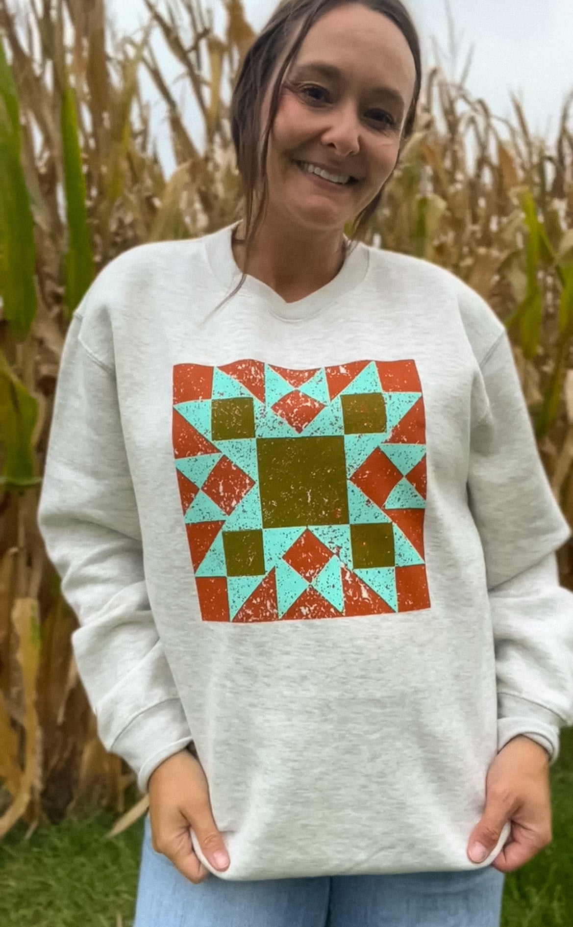 A woman wearing a light tan sweatshirt with a colorful geometric quilt-pattern graphic on the front, standing outdoors in front of a cornfield.