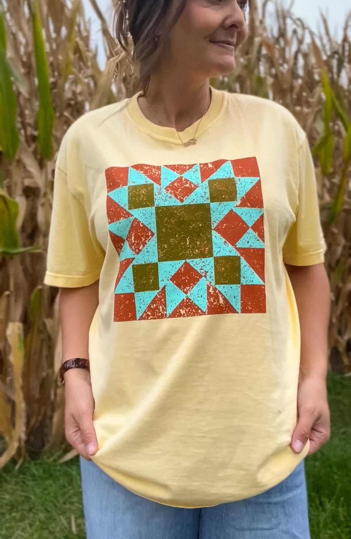 Model wearing butter yellow tee with a colorful barn quilt patter on the front, standing in front of a corn field. 