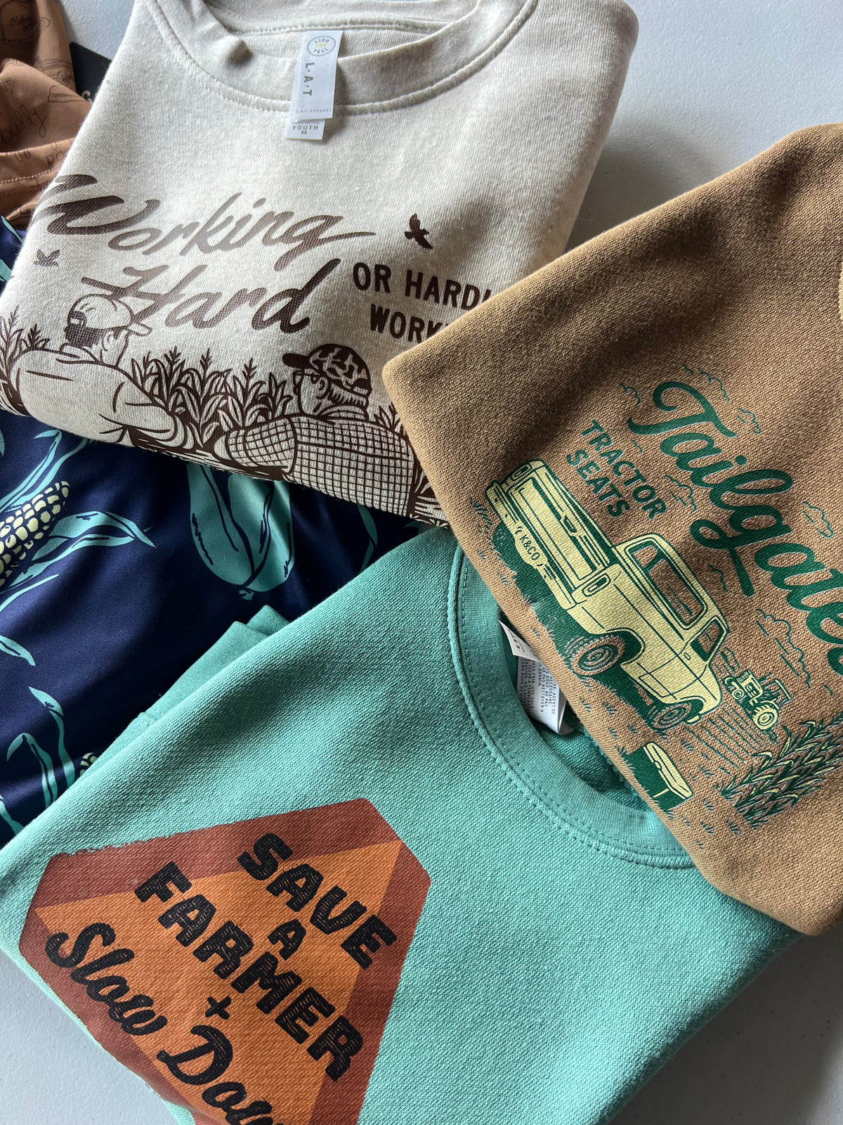 Close up picture of the working hard crewneck, tailgates and tractor seats crewneck, save a farmer crewneck, and shuck yeah polo. 
