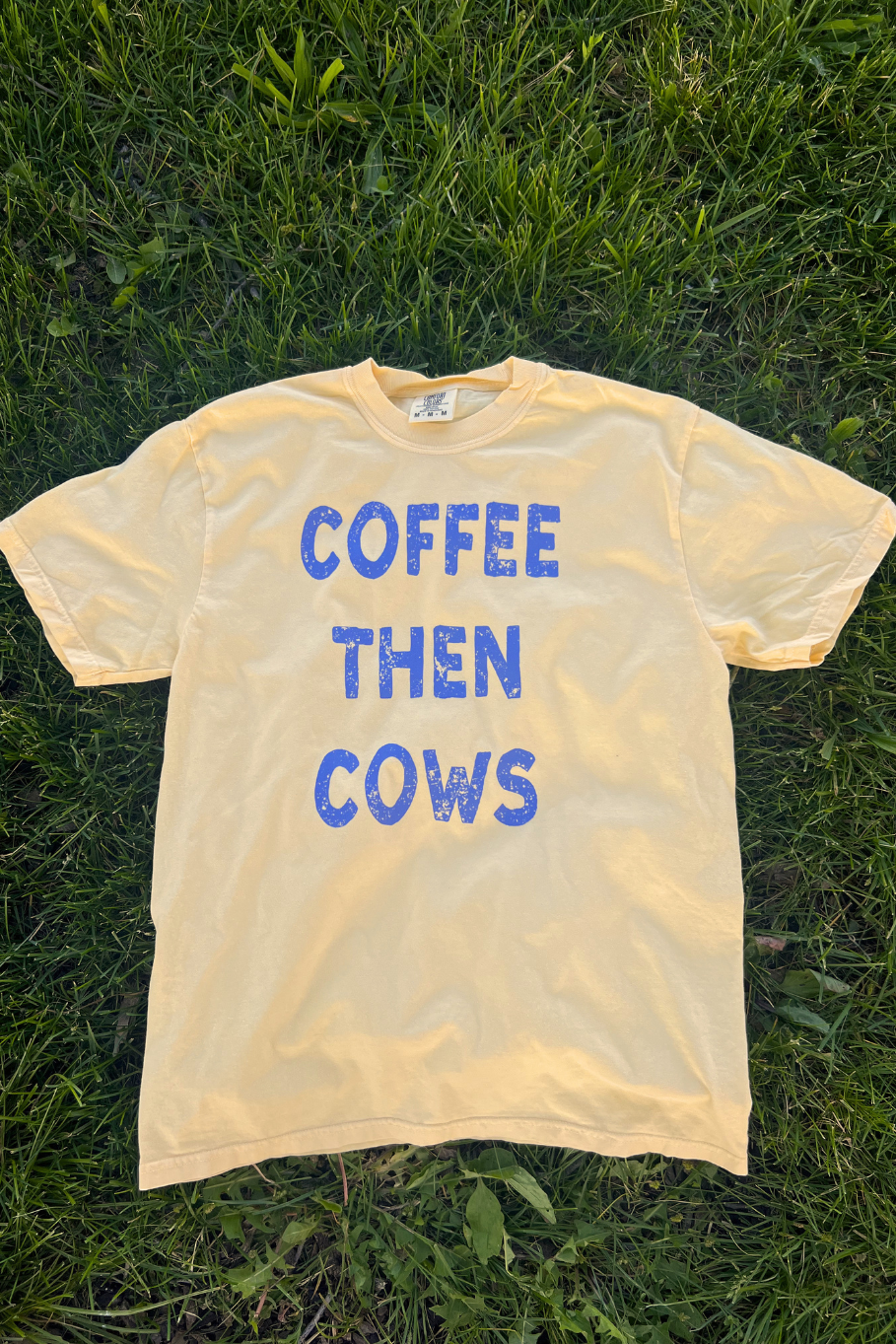 Butter yellow tee with blue text on the front of the tee "Coffee then cows" laying on grass.