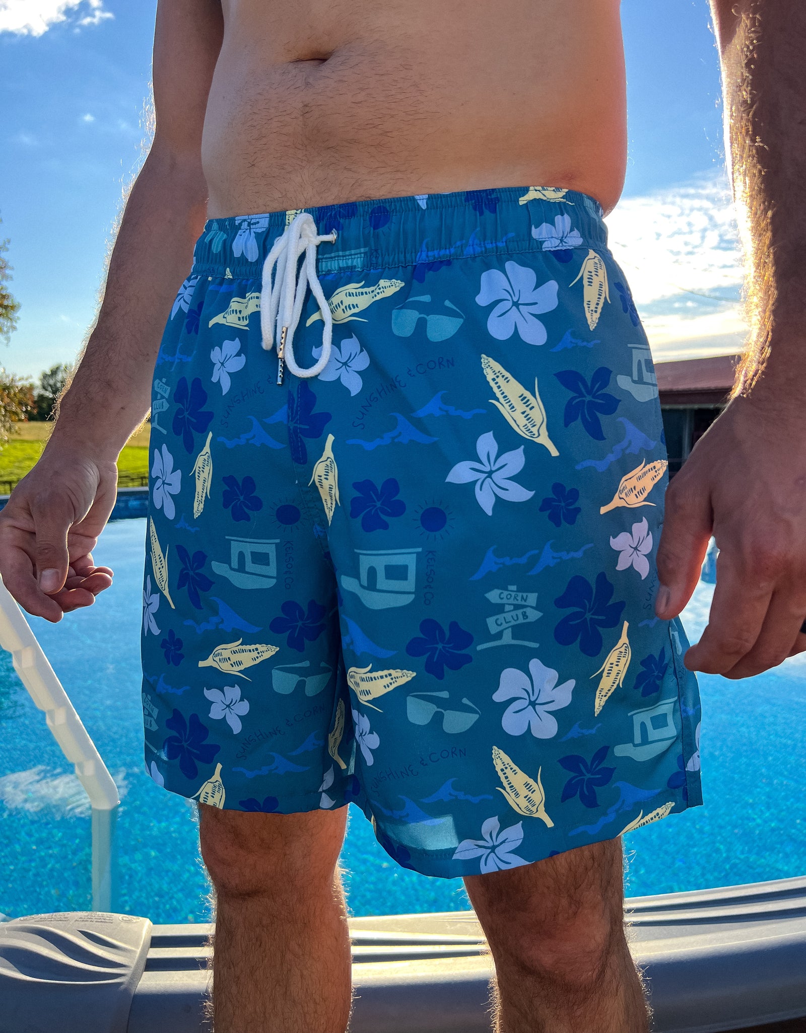 Man standing in front of pool wearing the corn club swim trunks - a Hawaiian inspired print with corn. 