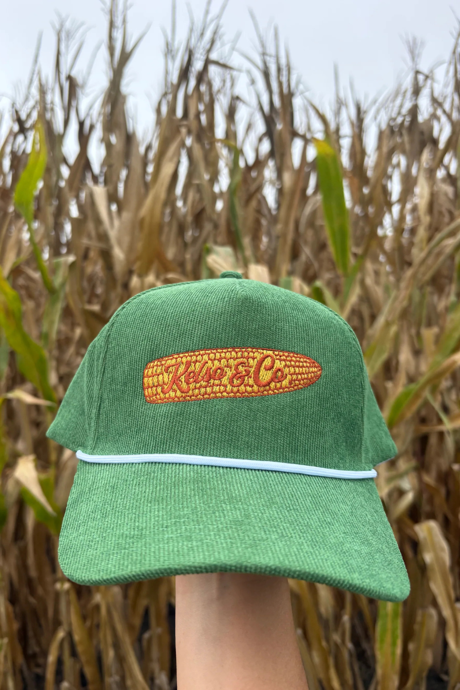 A green trucker hat with a white visor trim and a yellow embroidered corn patch reading 'Kelso & Co' is held up in front of a blurred cornfield.