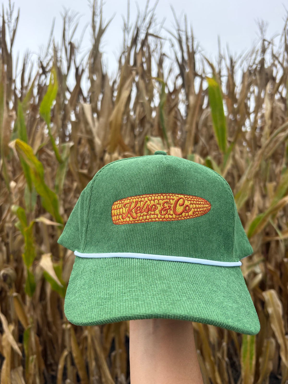 A green trucker hat with a white visor trim and a yellow embroidered corn patch reading 'Kelso &amp; Co' is held up in front of a blurred cornfield.