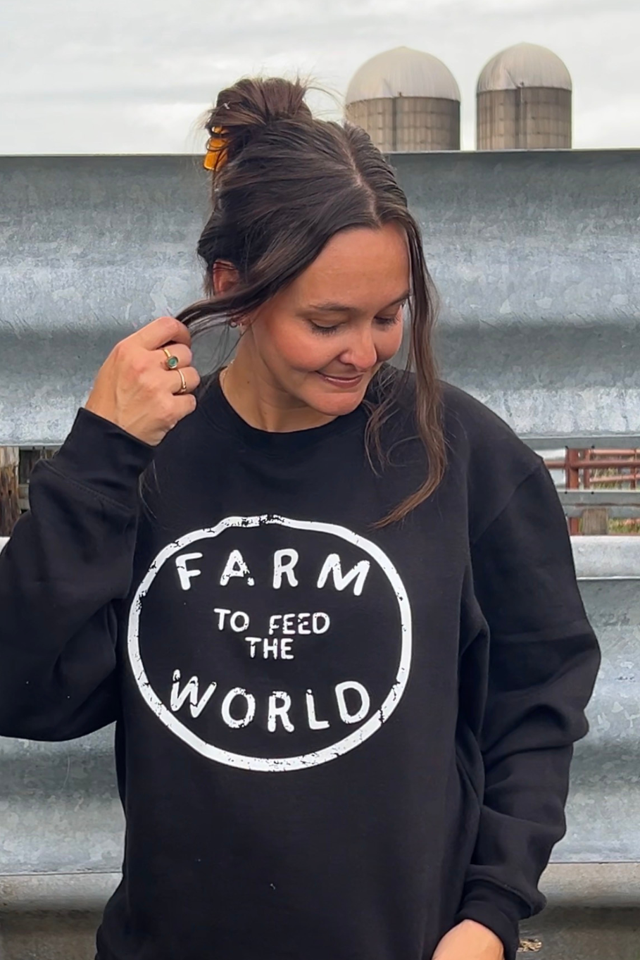 A woman wearing a black crewneck sweatshirt with a white distressed circular graphic that reads 'FARM TO FEED THE WORLD'. She is standing outdoors in front of a corrugated metal wall with silos in the background.