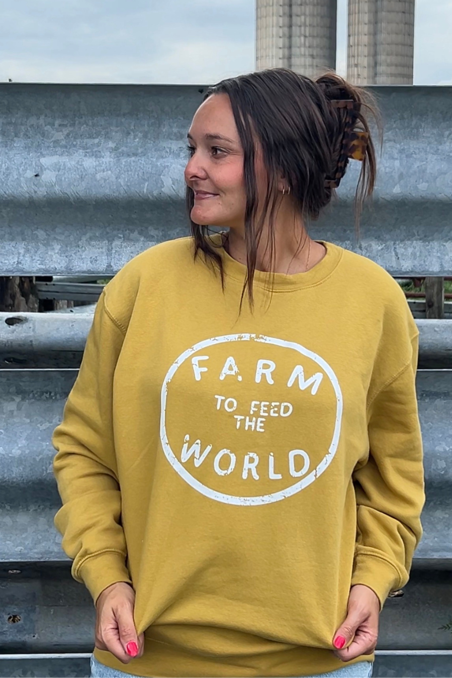 A woman wearing a mustard yellow crewneck sweatshirt with a white distressed graphic that reads 'FARM TO FEED THE WORLD', standing outdoors in front of a corrugated metal wall.