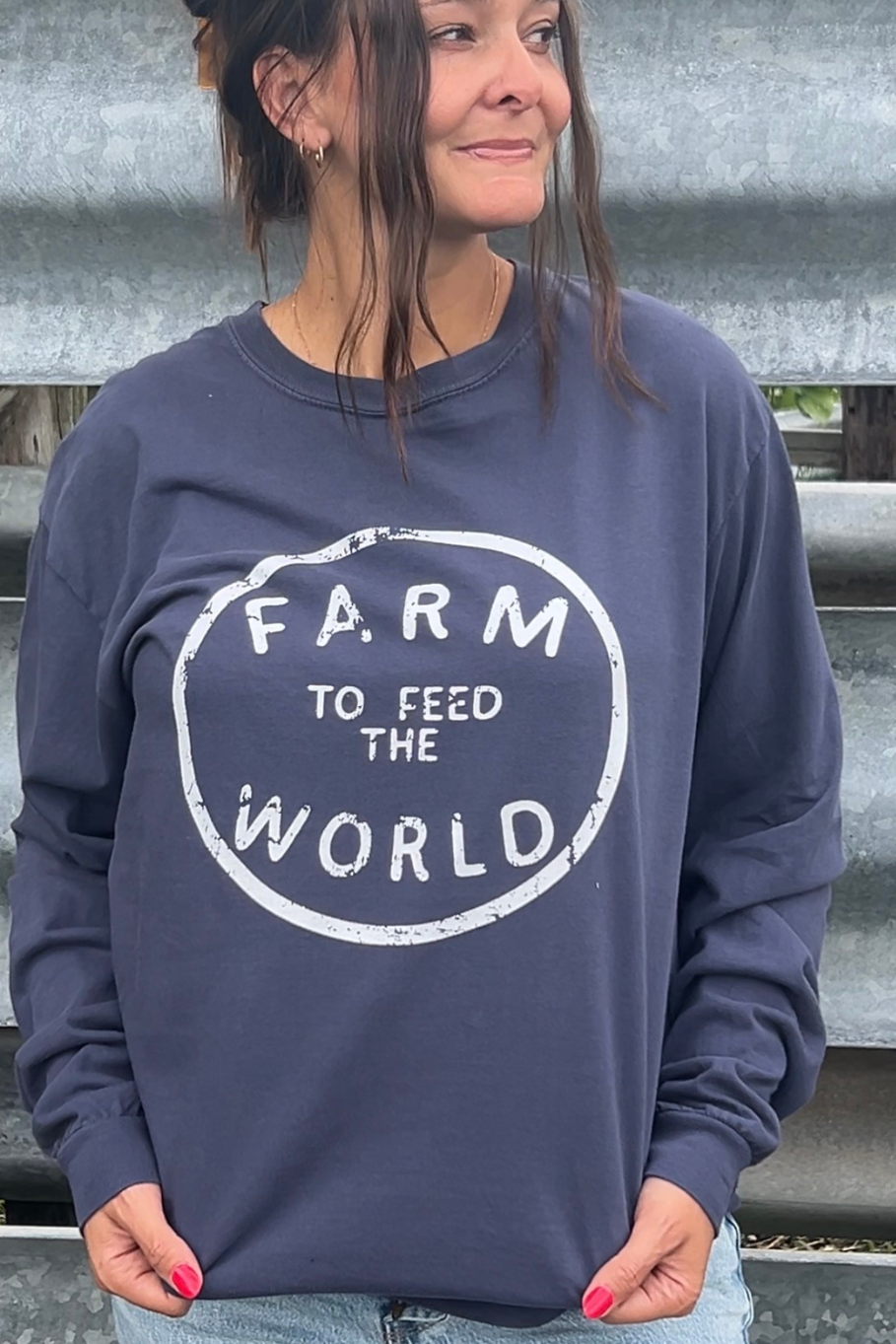 A woman wearing a navy blue crewneck sweatshirt with a distressed white graphic that reads 'FARM TO FEED THE WORLD' in a circle, standing in front of a gray metal back ground.