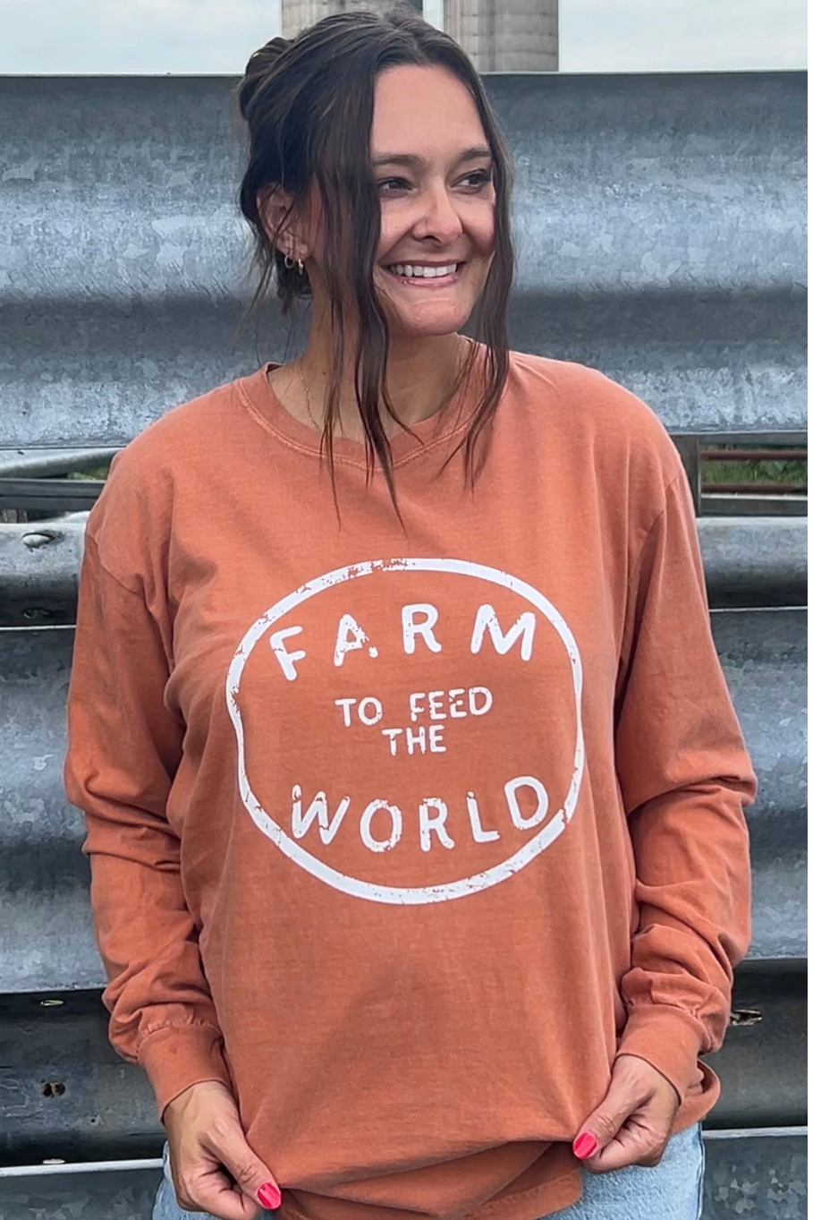 A woman wearing a burnt orange long-sleeve crewneck sweatshirt with a white distressed graphic that reads 'FARM TO FEED THE WORLD'. She is standing outdoors in front of corrugated metal siding.