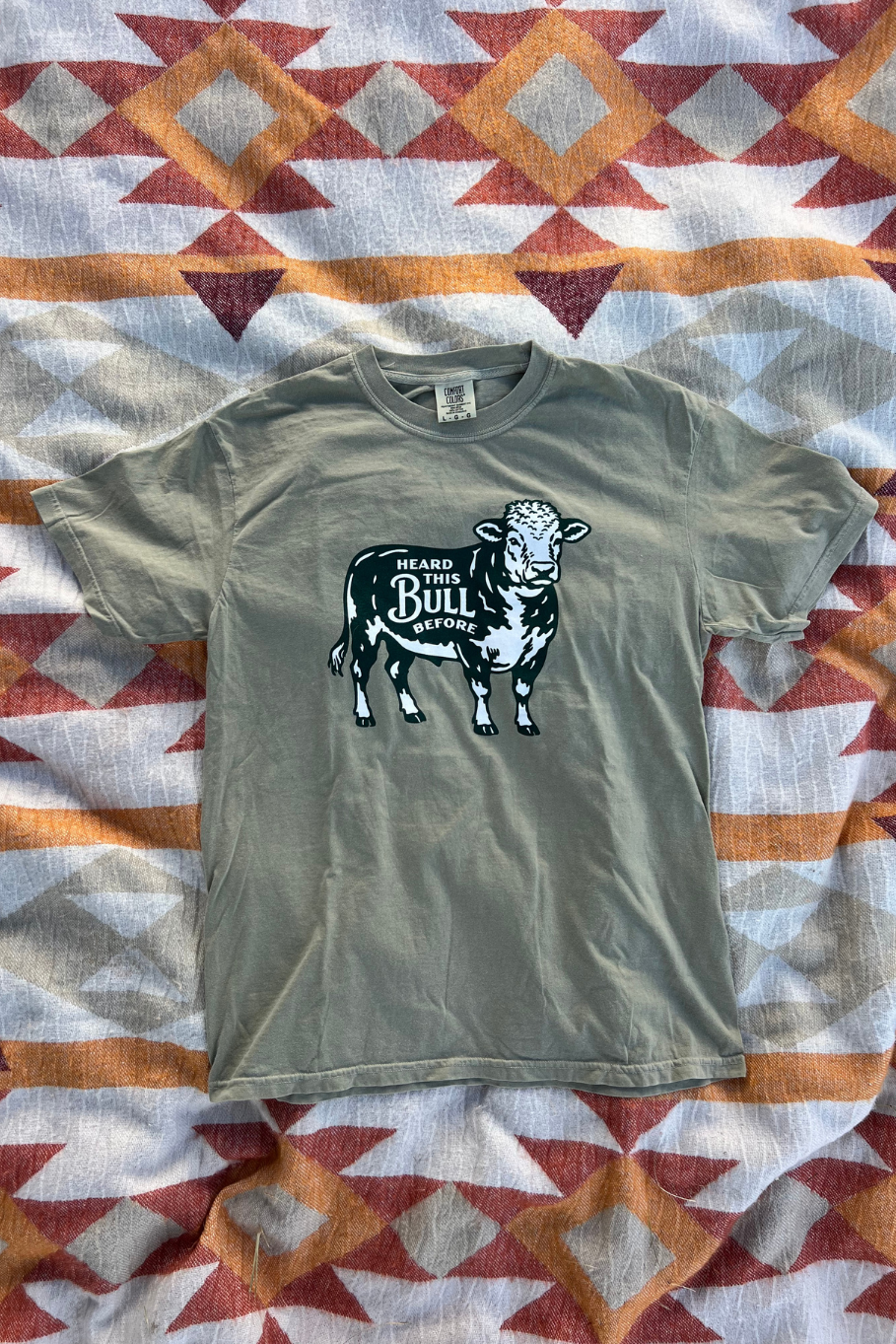 A short-sleeve crewneck t-shirt in khaki, laid flat on a patterned quilt. The shirt features a graphic of a bull with the text 'Heard that Bull Before' printed on the front. 