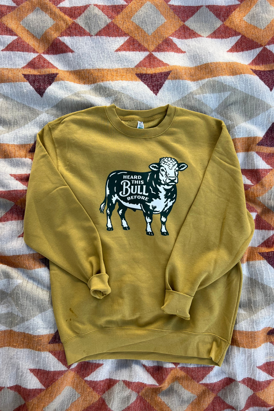 Flat lay picture of an antique gold colored crewneck with a bull graphic on the front that says heard this bull before