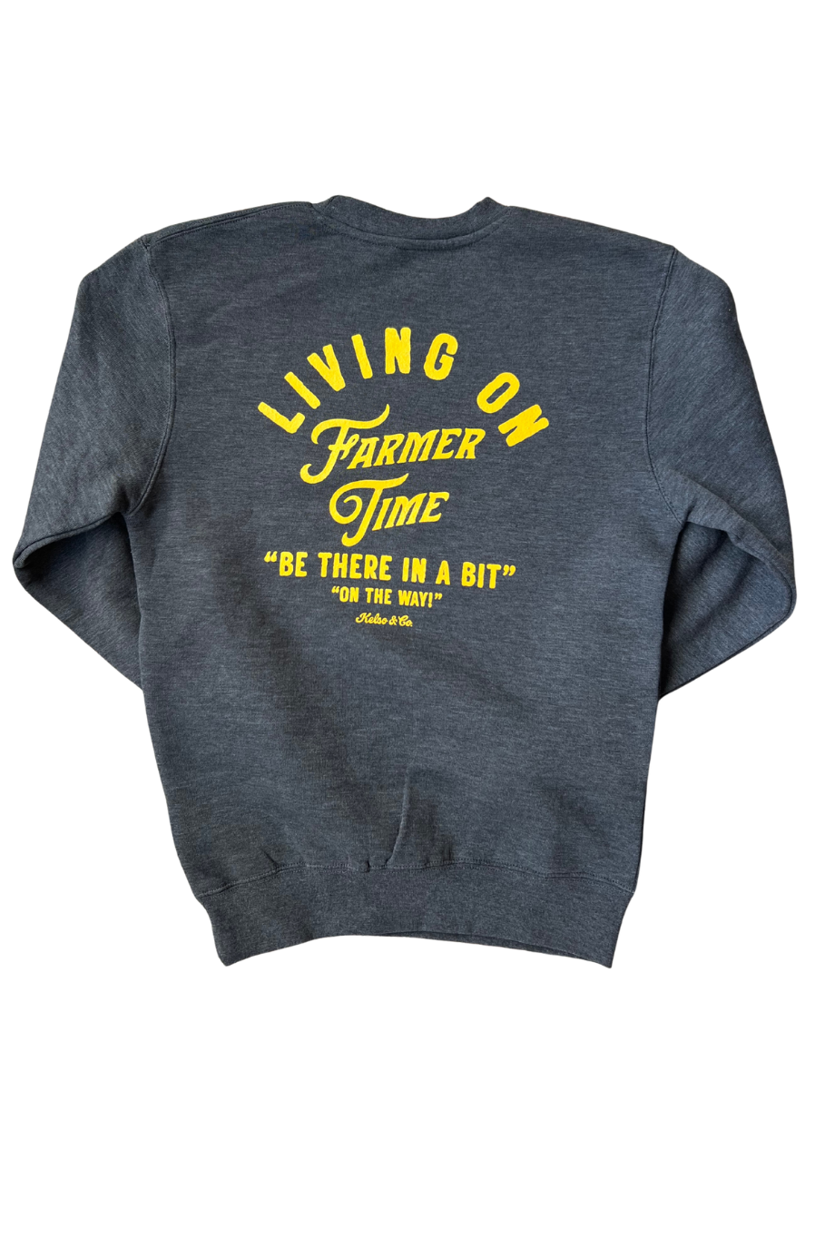A dark heather gray sweatshirt shown from the back, featuring a bold yellow graphic print that reads 'LIVING ON FARMER TIME' and 'BE THERE IN A BIT ON THE WAY'.