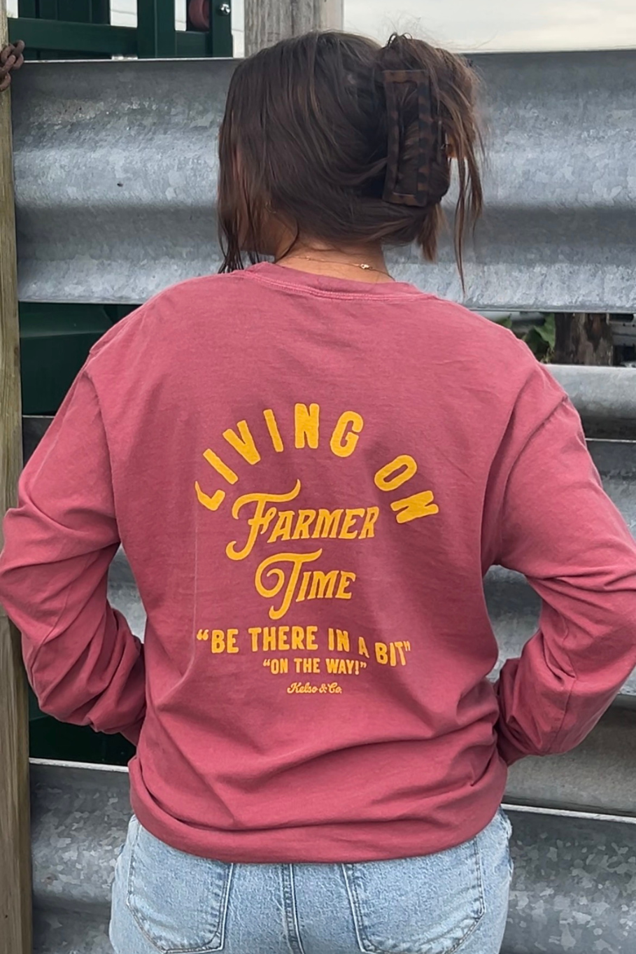 A person viewed from behind wearing a brick colored long-sleeve graphic sweatshirt with yellow text that reads 'LIVING ON FARMER TIME' and 'BE THERE IN A BIT ON THE WAY'.