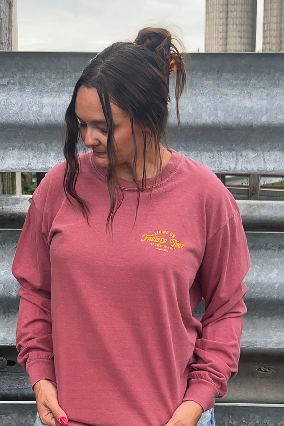 A woman wearing a brick colored long-sleeve crewneck sweatshirt with a yellow graphic on the left chest, standing in front of a corrugated metal wall.