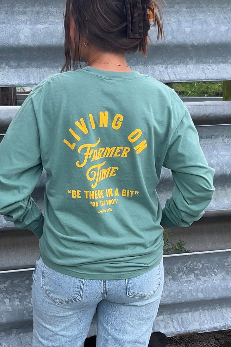 A person viewed from behind wearing a sage green long-sleeve graphic shirt with yellow text reading 'LIVING ON Farmer Time' and 'BE THERE IN A BIT' on the back, paired with light blue jeans in front of a corrugated metal wall.