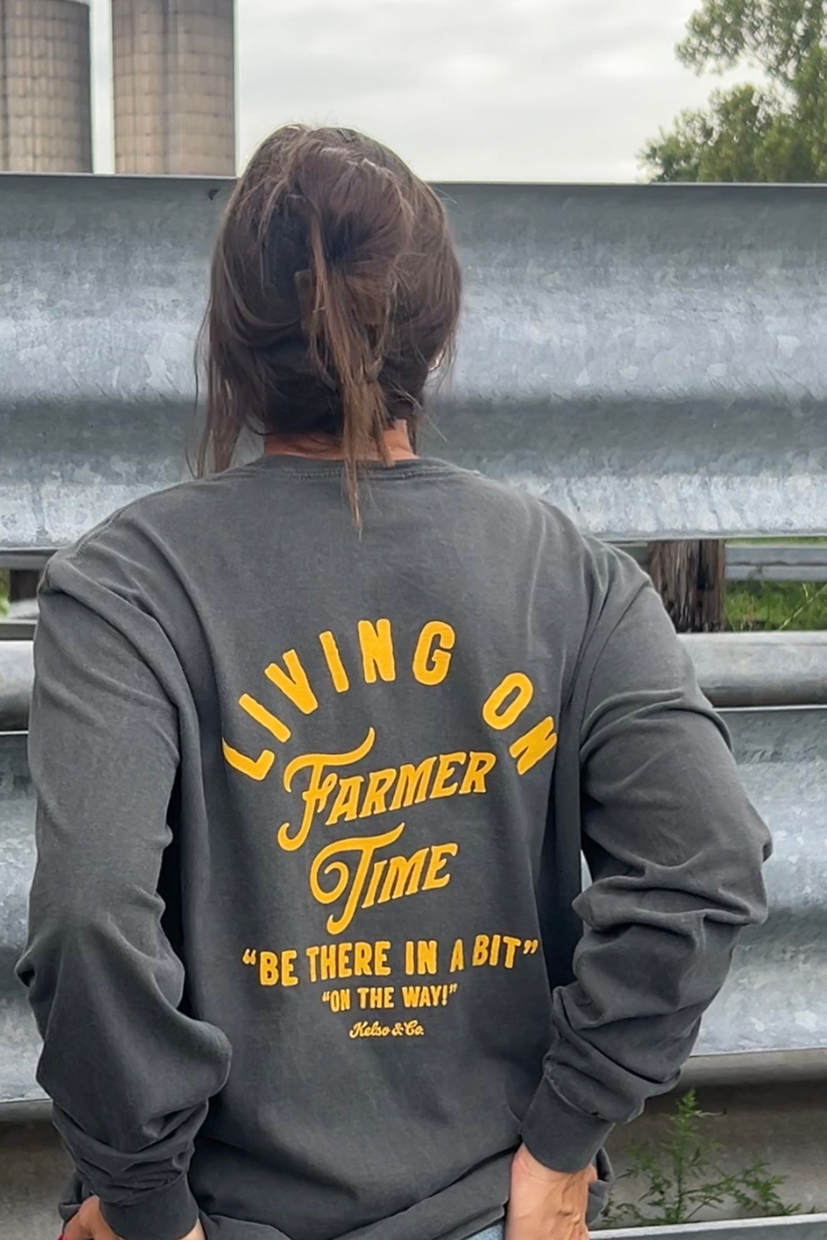 A person viewed from behind wearing a dark gray long-sleeve sweatshirt with a large yellow graphic text on the back reading 'LIVING ON Farmer Time' and 'BE THERE IN A BIT' 'ON THE WAY!'.