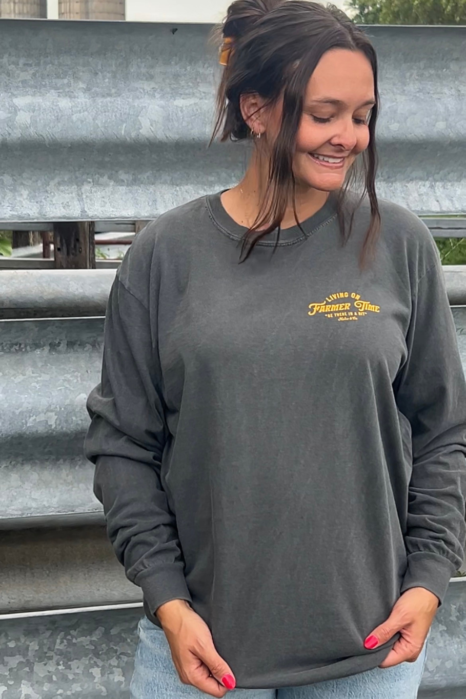 A woman wearing a dark charcoal gray long-sleeve t-shirt with a yellow embroidered logo on the left chest, standing in front of corrugated metal siding.