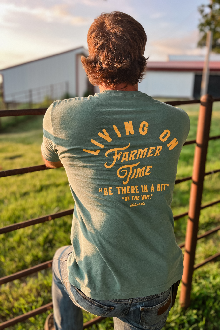 Guy leaning on a fence with a tshirt on that says "living on farmer time, be there in a bit"