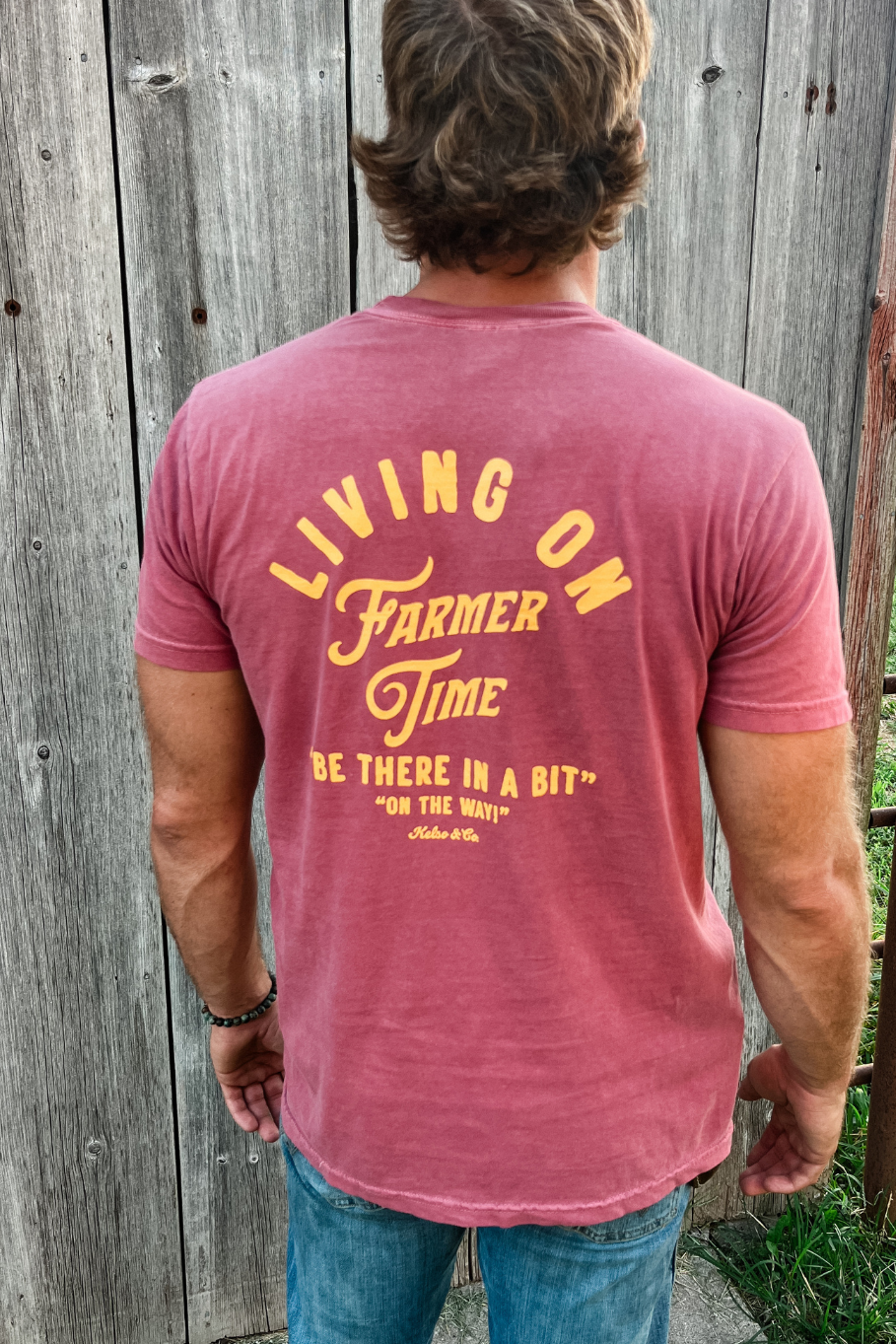 man in front of a barn wearing a graphic tshirt that says on the back &quot;Living on Farmer Time, Be there in a bit&quot;